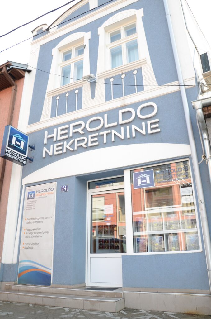 Blue building with "HEROLDO NEKRETNINE" sign and logo. Glass door and windows display property ads. White accents.